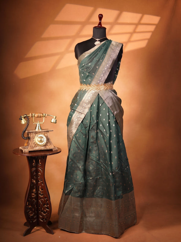 A dark teal green saree with silver zari embroidery and a wide metallic silver border, displayed on a black mannequin. The mannequin is set against a warm brown backdrop with decorative window shadow overlays, and a vintage wooden rotary telephone sits on a carved side table to the left.