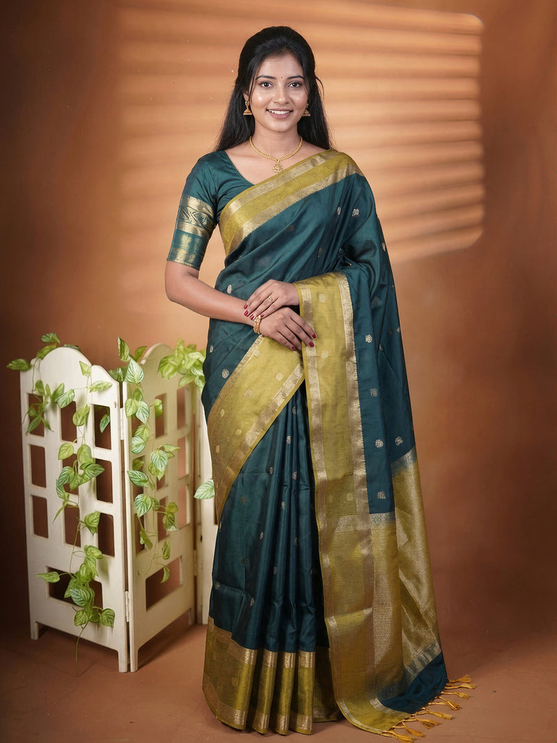 A woman modeling a dark teal green and gold zari saree with a matching blouse, standing in front of a warm brown studio backdrop with decorative leafy props on the left.