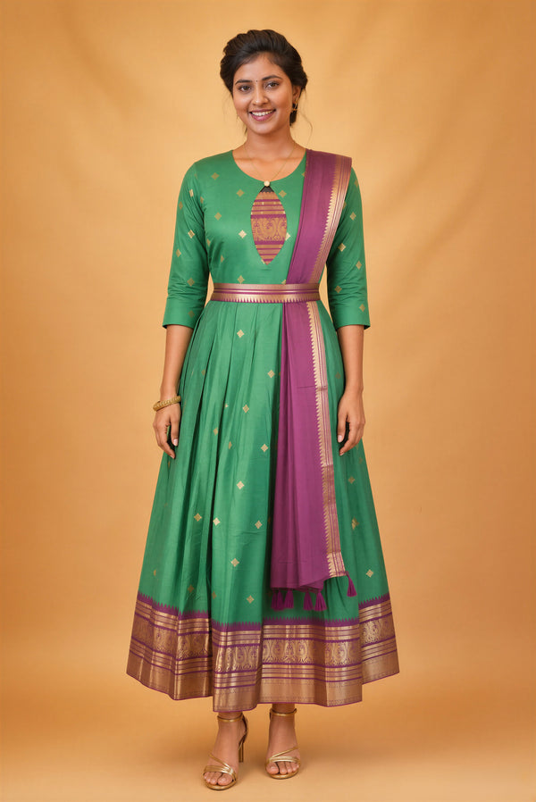 A woman modeling a green traditional Anarkali gown with gold motifs, a purple border, and a matching purple dupatta, set against a solid warm beige studio backdrop.