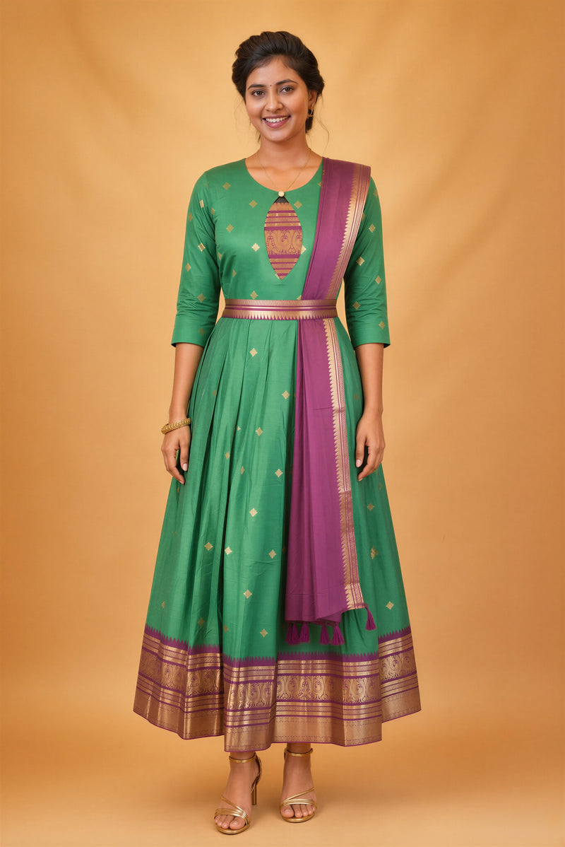 A woman modeling a green traditional Anarkali gown with gold motifs, a purple border, and a matching purple dupatta, set against a solid warm beige studio backdrop.