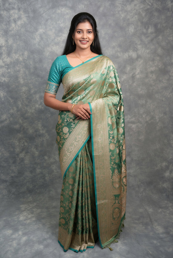 A woman modeling a traditional Indian saree with a teal green blouse and a sage green and gold patterned saree, standing against a textured gray studio backdrop.