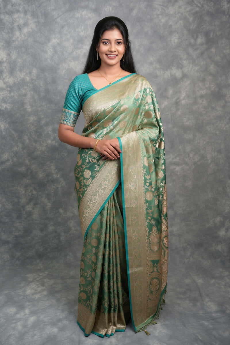 A woman modeling a traditional Indian saree with a teal green blouse and a sage green and gold patterned saree, standing against a textured gray studio backdrop.