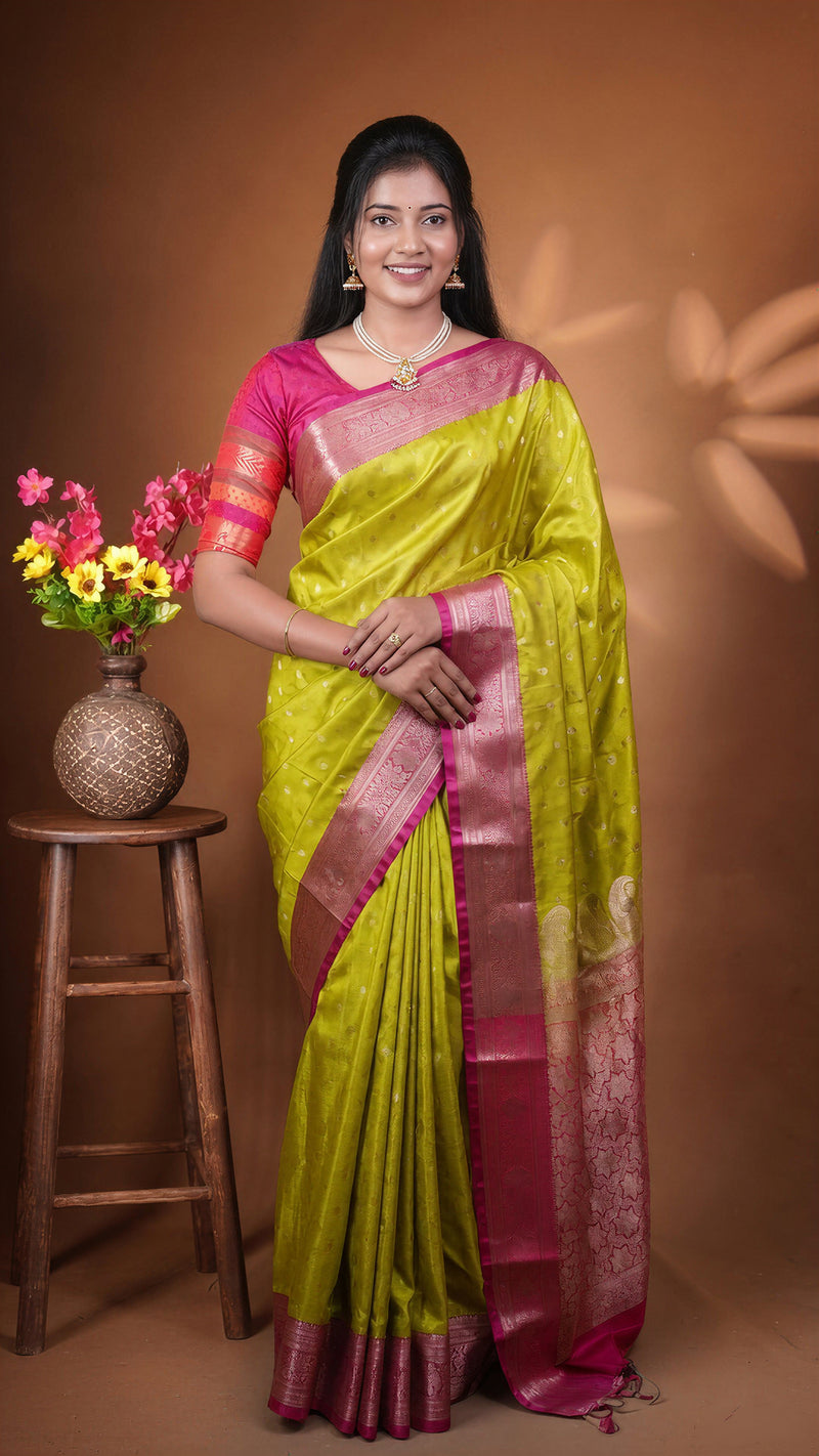 A woman wearing a bright yellow-green and magenta saree stands against a warm brown studio backdrop, with a small wooden stool and vase of flowers to her left.