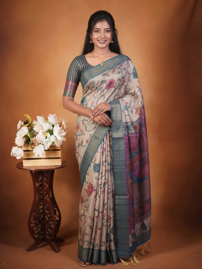A woman modeling a traditional Indian saree with a floral print, standing against a warm brown studio backdrop next to a small wooden table with white flowers.