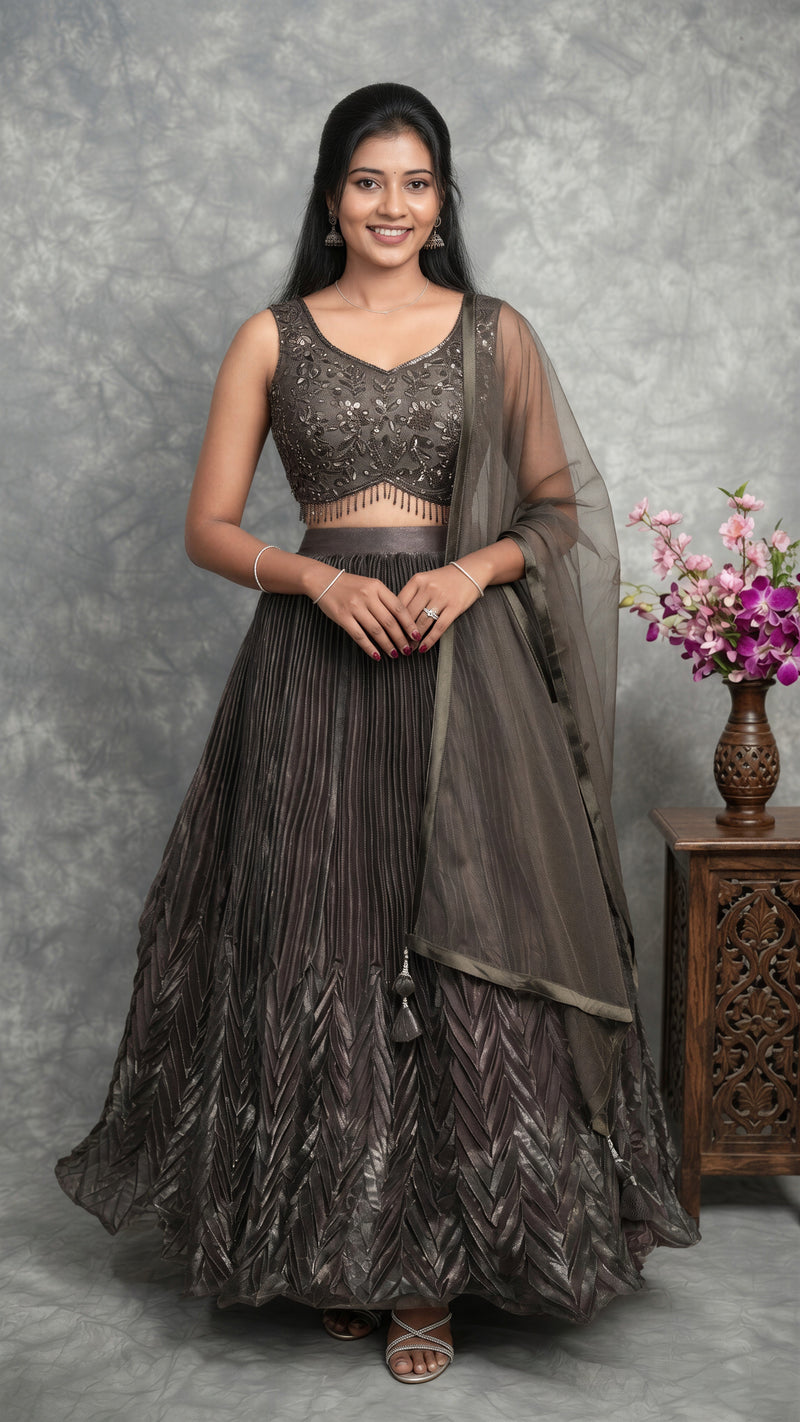 A woman modeling a dark brown embroidered traditional Indian outfit with a pleated skirt, embroidered crop top, and sheer dupatta, standing against a textured gray backdrop.