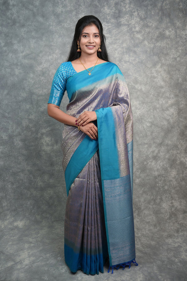 A woman modeling a blue and silver gradient silk saree with a matching blue embroidered blouse, standing against a textured gray backdrop.