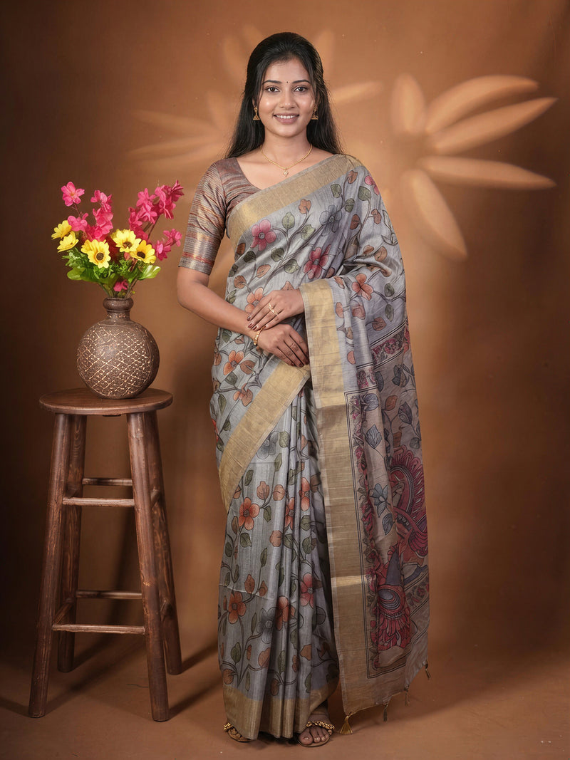 A woman wearing a gray and gold floral saree stands next to a wooden stool with a vase of colorful flowers, set against a warm brown backdrop with leaf shadows.