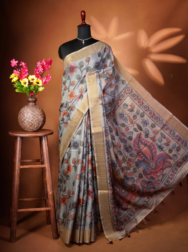 A gray silk saree with a floral print and a wide beige border draped on a black mannequin, styled in a studio setting with a wooden stool holding a vase of pink and yellow flowers against a warm brown background.