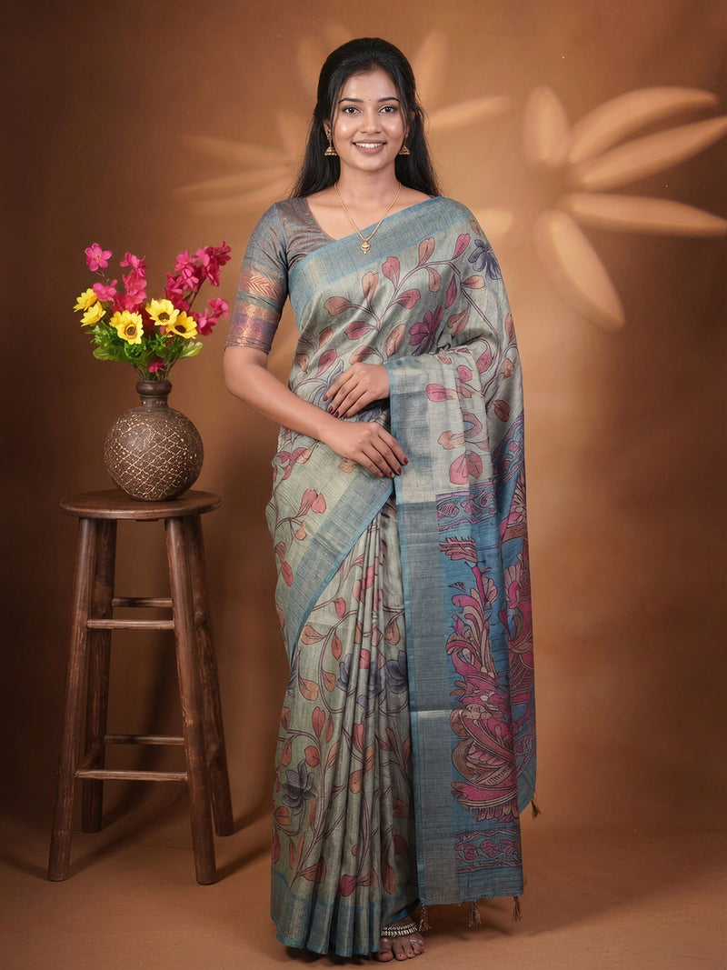 A woman modeling a light blue-gray saree with pink and green floral patterns, standing in front of a warm brown backdrop. A wooden stool with a vase of colorful flowers is on the left.