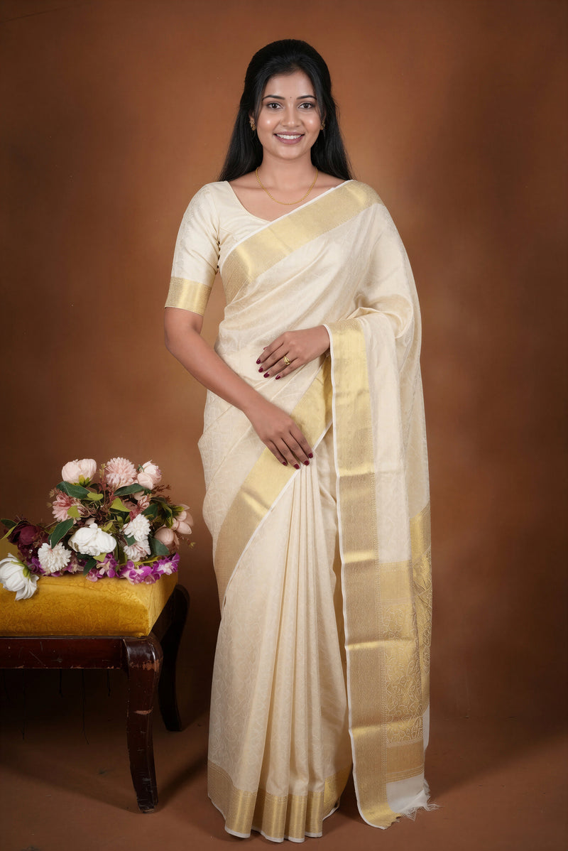 A woman modeling a cream-colored silk saree with a wide gold border, standing against a solid brown studio backdrop. A small yellow bench with flowers is visible on the left side of the frame.