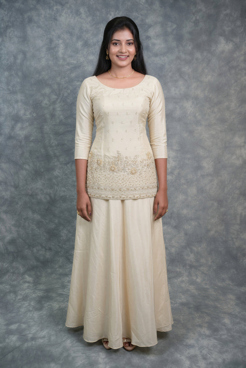 A woman modeling a cream-colored traditional Indian outfit consisting of a three-quarter sleeve kurta with floral embroidery and a matching long skirt, set against a mottled gray studio backdrop.