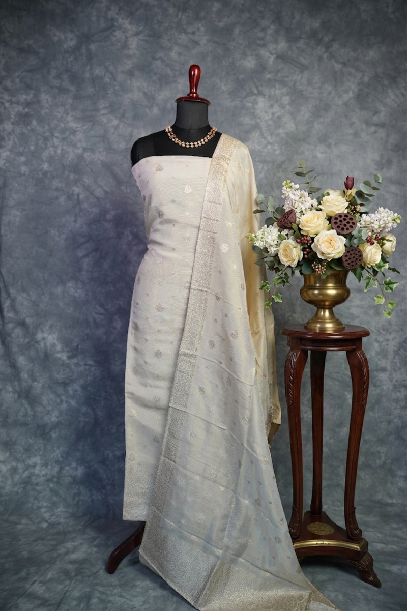 White saree with gold border on a mannequin, next to a small table with flowers against a gray background