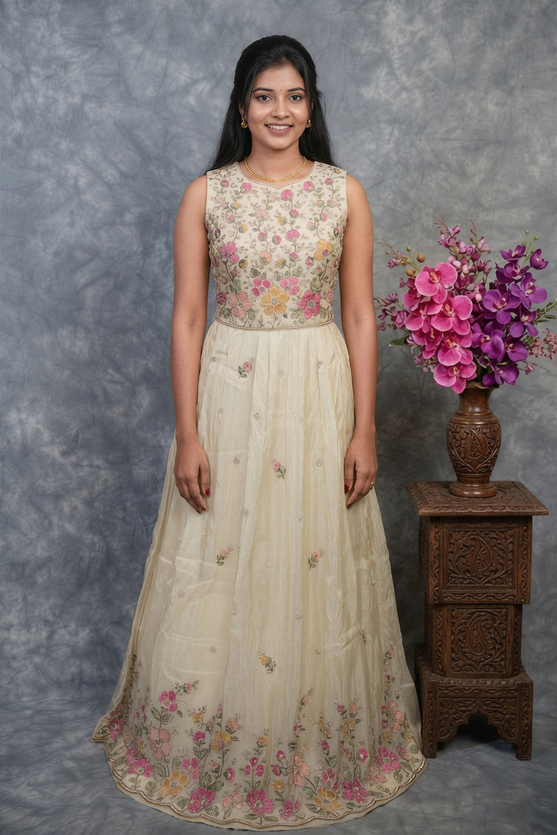 A woman modeling a sleeveless cream-colored traditional gown with pink and green floral embroidery on the bodice and hem, standing against a textured gray backdrop next to a wooden side table with a vase of purple flowers.