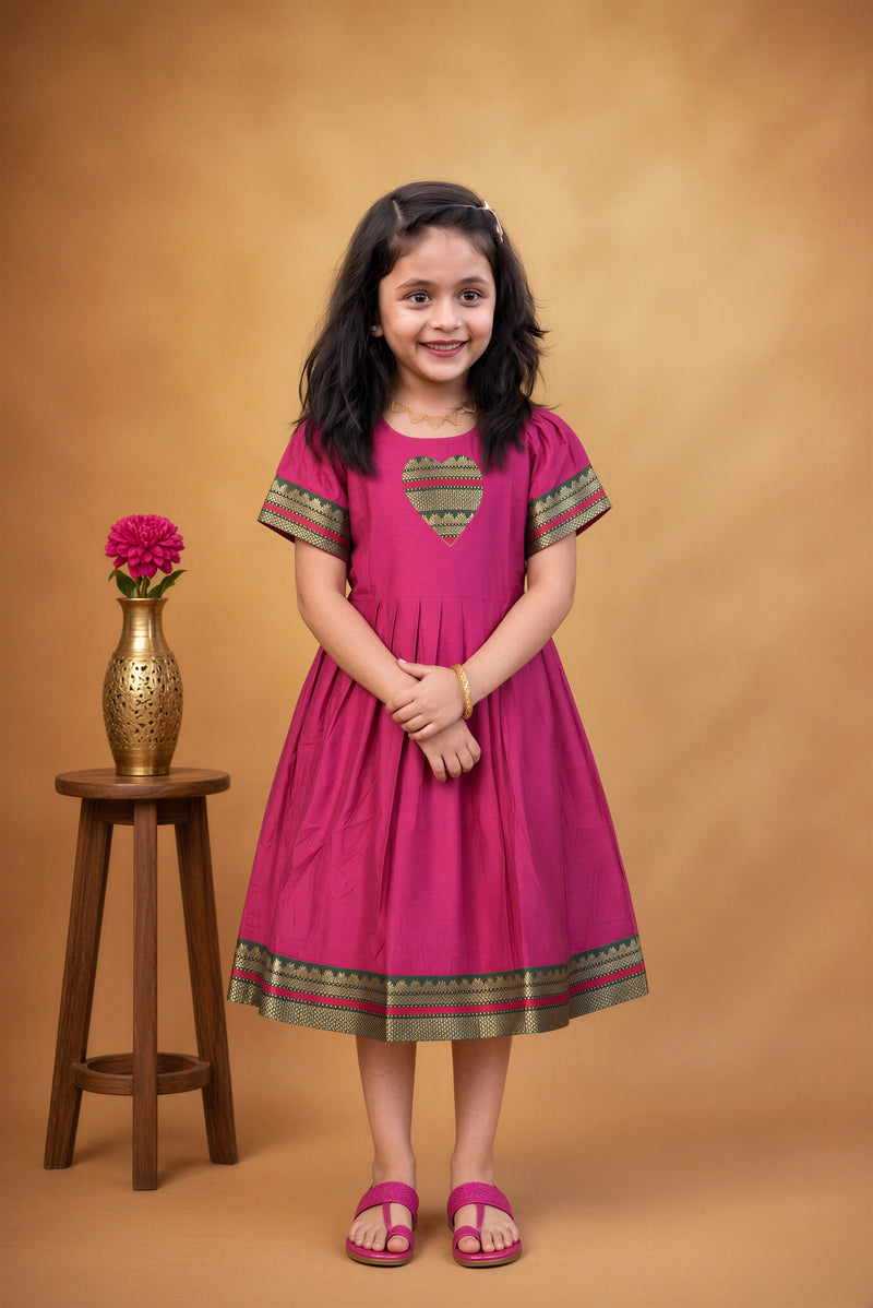 A young girl stands in a studio wearing a bright pink dress with green and gold patterned trim on the chest and hem. She is posed next to a small wooden stool holding a gold vase with a pink flower, against a warm brown backdrop.