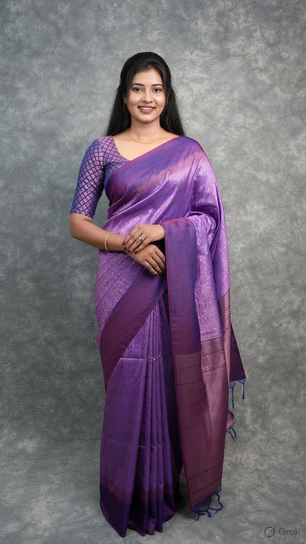 A woman modeling a purple silk saree with a matching purple patterned blouse, standing against a textured gray studio backdrop.