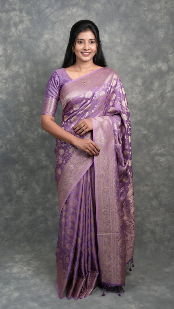 A woman modeling a lavender purple silk saree with gold zari floral patterns and a matching purple blouse, standing against a textured gray studio backdrop.