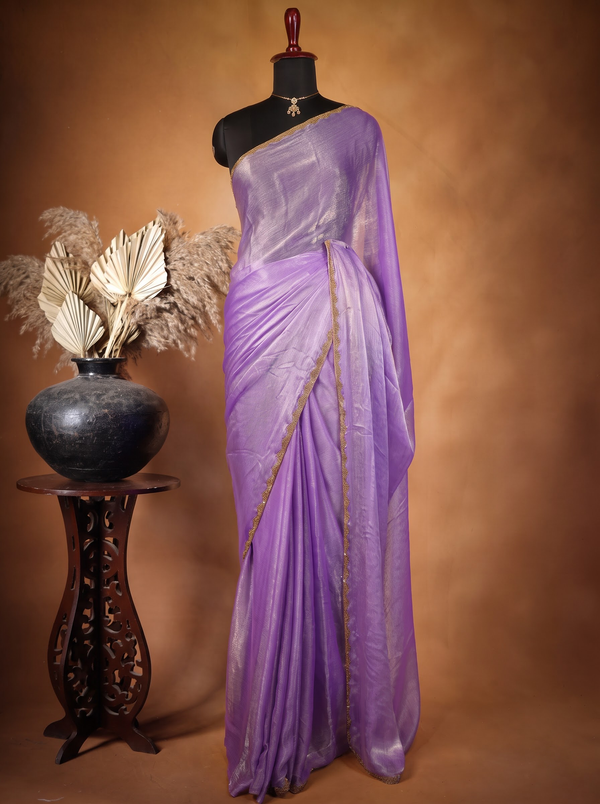 A lavender purple saree with gold zari embroidery draped on a black mannequin torso, set against a warm brown studio backdrop. A carved wooden stand holding a dark vase with dried pampas grass is positioned to the left of the mannequin.
