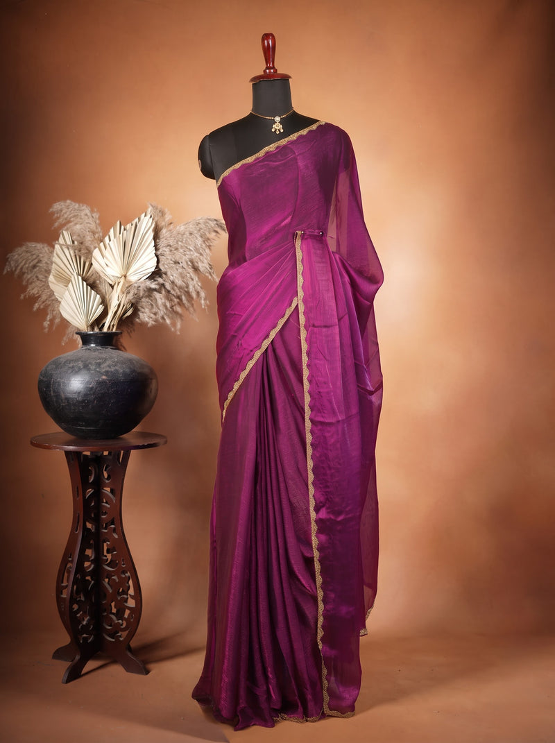 A deep purple silk saree with a gold border draped on a black mannequin, set against a warm brown studio backdrop. A small carved wooden stand holding a black vase with dried pampas grass is positioned to the left.