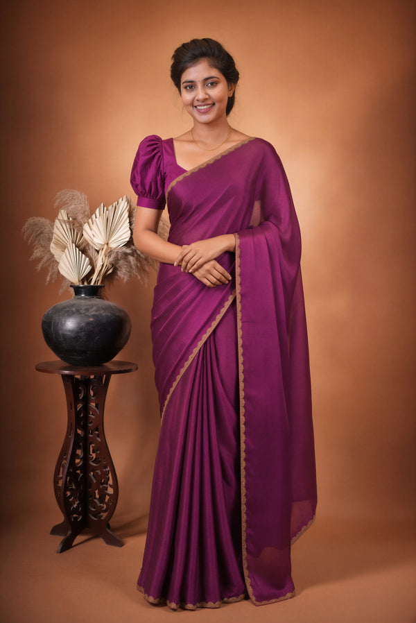 A woman wearing a deep purple silk saree with gold border detailing, standing in front of a warm brown studio backdrop next to a decorative black vase on a wooden stand.