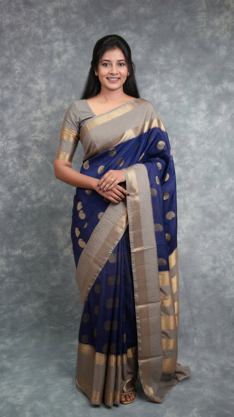 A woman modeling a traditional Indian saree with a deep blue body, beige border, and gold motifs, standing against a textured gray backdrop.