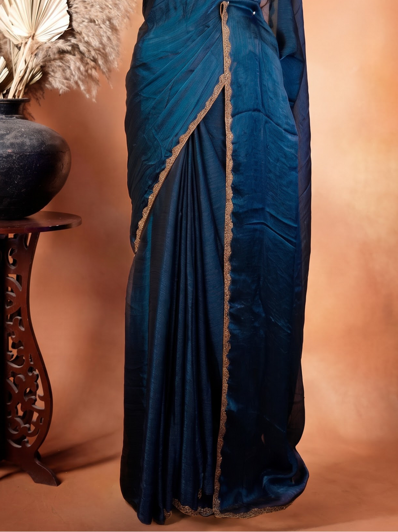 A dark teal blue silk saree with gold border trim draped on a black mannequin, set against a warm brown studio backdrop with a decorative ceramic pot and dried palm leaves on a wooden stand.