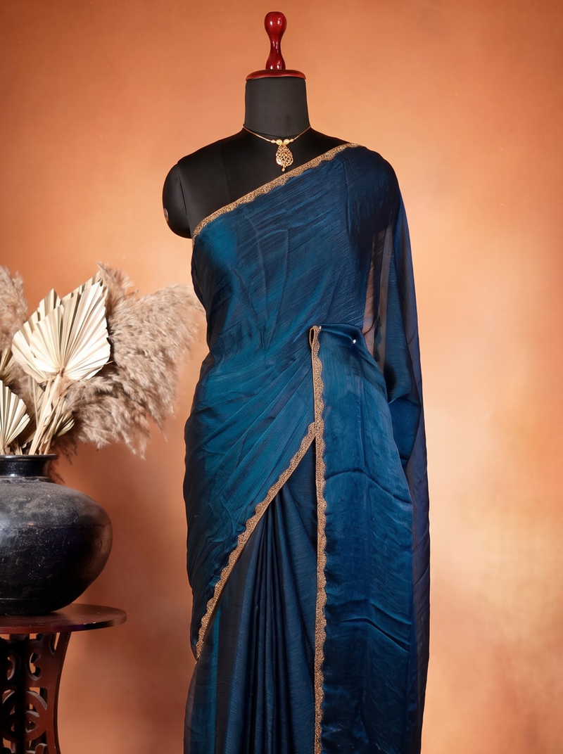 A dark teal blue silk saree with gold border trim draped on a black mannequin, set against a warm brown studio backdrop with a decorative ceramic pot and dried palm leaves on a wooden stand.
