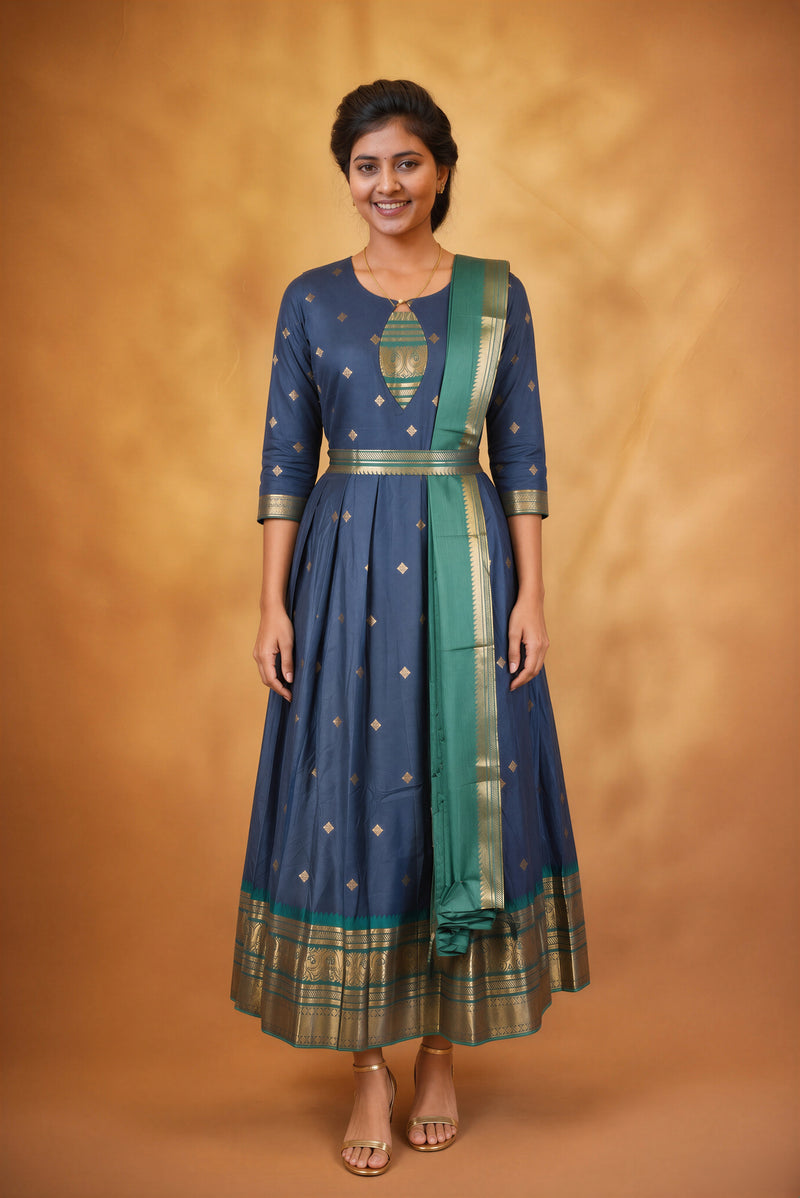A woman modeling a navy blue traditional Anarkali gown with gold zari embroidery, a green border, and a matching green dupatta, set against a warm brown studio backdrop.