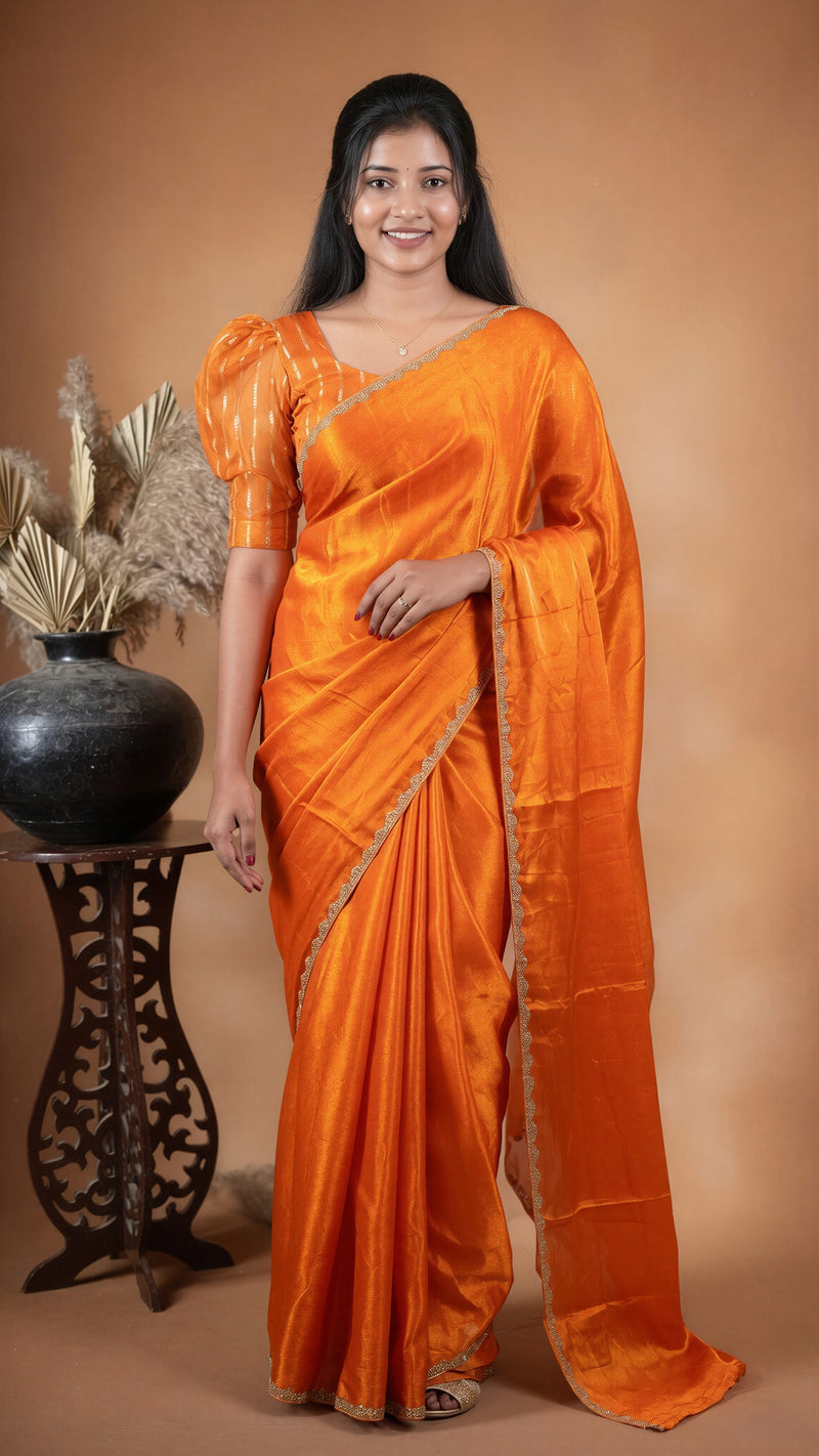 A woman modeling a bright orange silk saree with a matching blouse, standing against a warm brown studio backdrop next to a decorative black vase.