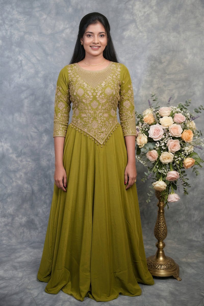 A woman modeling a long olive green traditional gown with beige floral embroidery on the bodice and sleeves, standing against a mottled gray backdrop next to a floral arrangement.