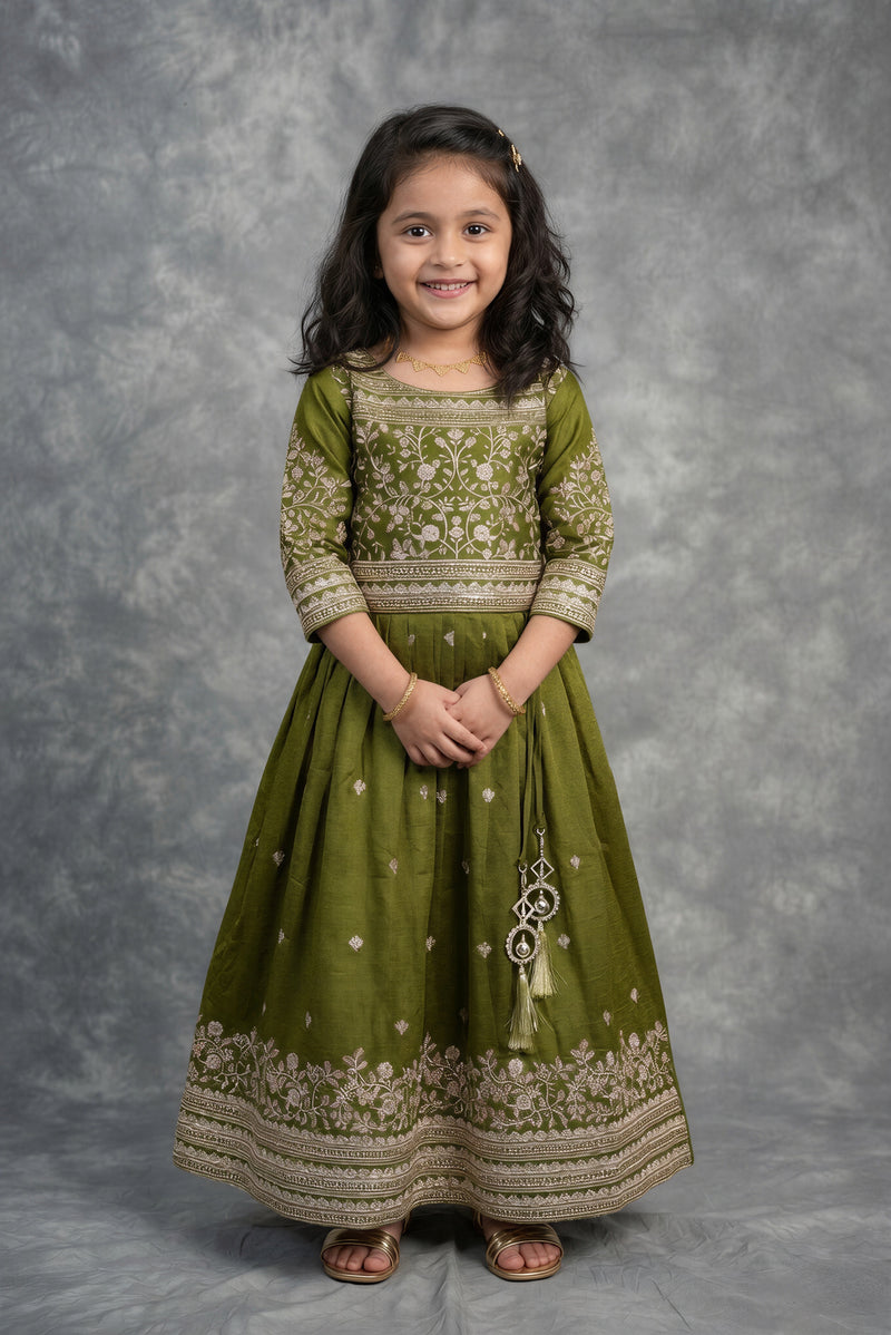 A young girl with dark hair stands against a textured gray studio backdrop wearing an olive green traditional dress with beige floral embroidery and gold detailing.