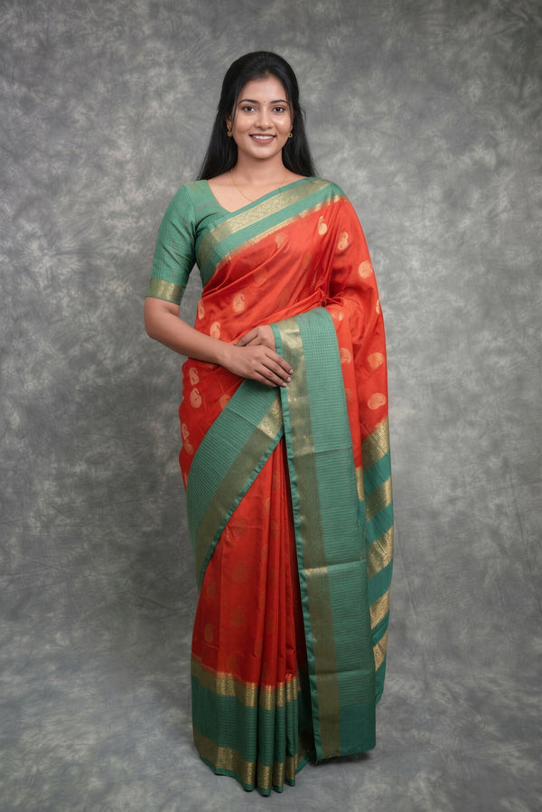 A woman modeling a traditional Indian saree with a red body, green border and blouse, decorated with gold zari motifs, set against a textured gray studio backdrop.