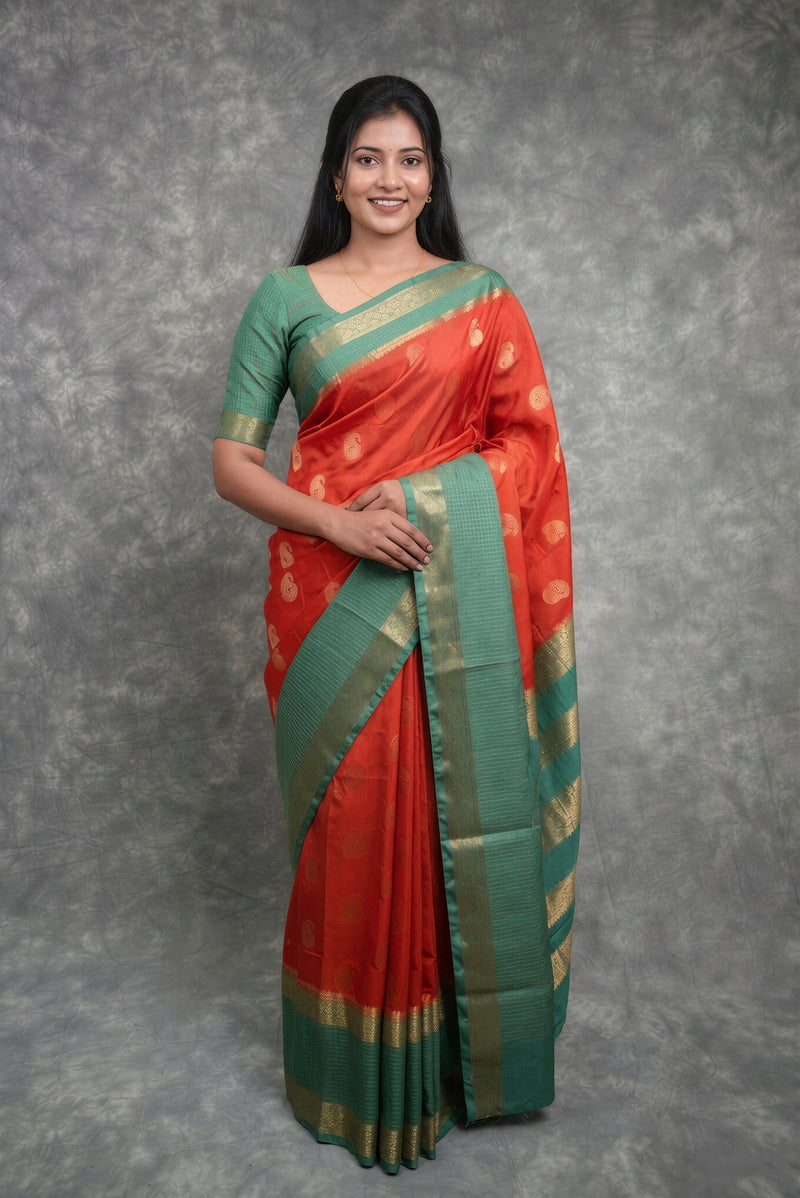 A woman modeling a traditional Indian saree with a red body, green border and blouse, decorated with gold zari motifs, set against a textured gray studio backdrop.
