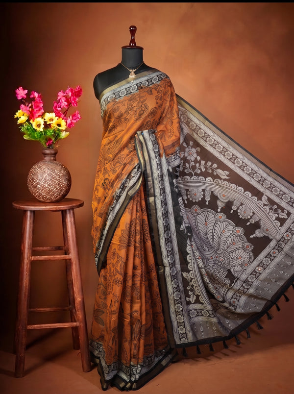 A Orange and black printed saree with a gray and white border, displayed on a black mannequin torso. The mannequin is set against a warm brown studio backdrop and is styled next to a wooden stool holding a vase of colorful flowers.