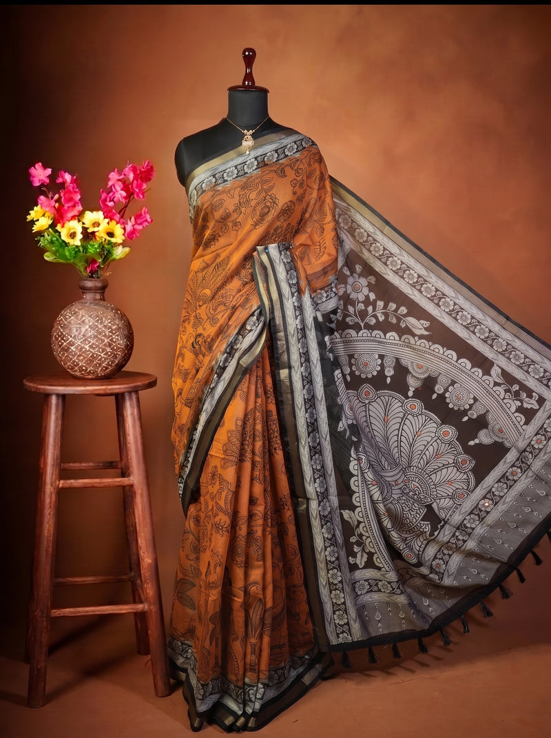 A Orange and black printed saree with a gray and white border, displayed on a black mannequin torso. The mannequin is set against a warm brown studio backdrop and is styled next to a wooden stool holding a vase of colorful flowers.