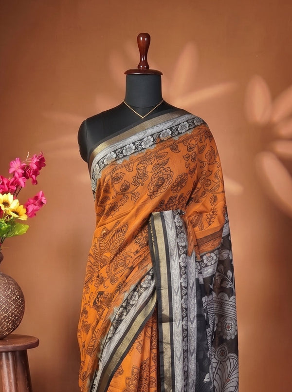 An orange and black printed saree draped on a black mannequin torso, styled next to a rustic wooden stool holding a vase of pink and yellow flowers against a warm brown backdrop.