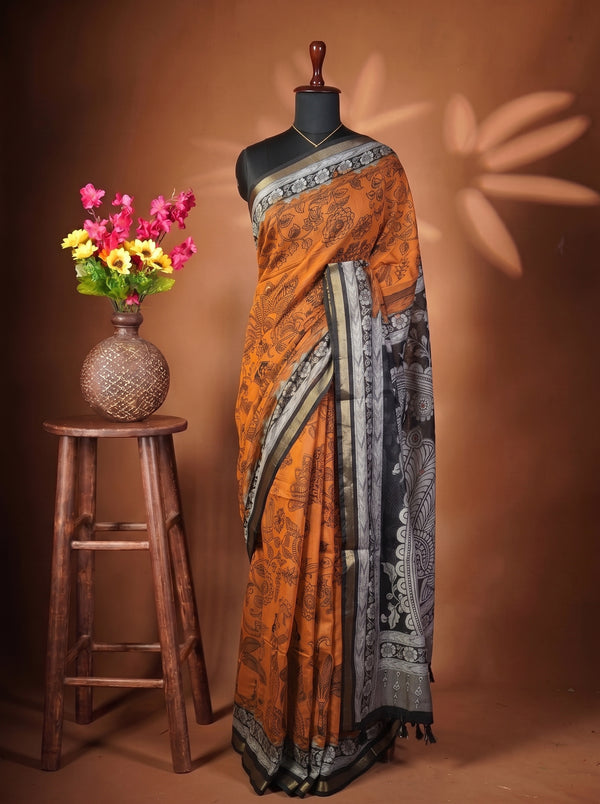An orange and black printed saree draped on a black mannequin torso, styled next to a rustic wooden stool holding a vase of pink and yellow flowers against a warm brown backdrop.