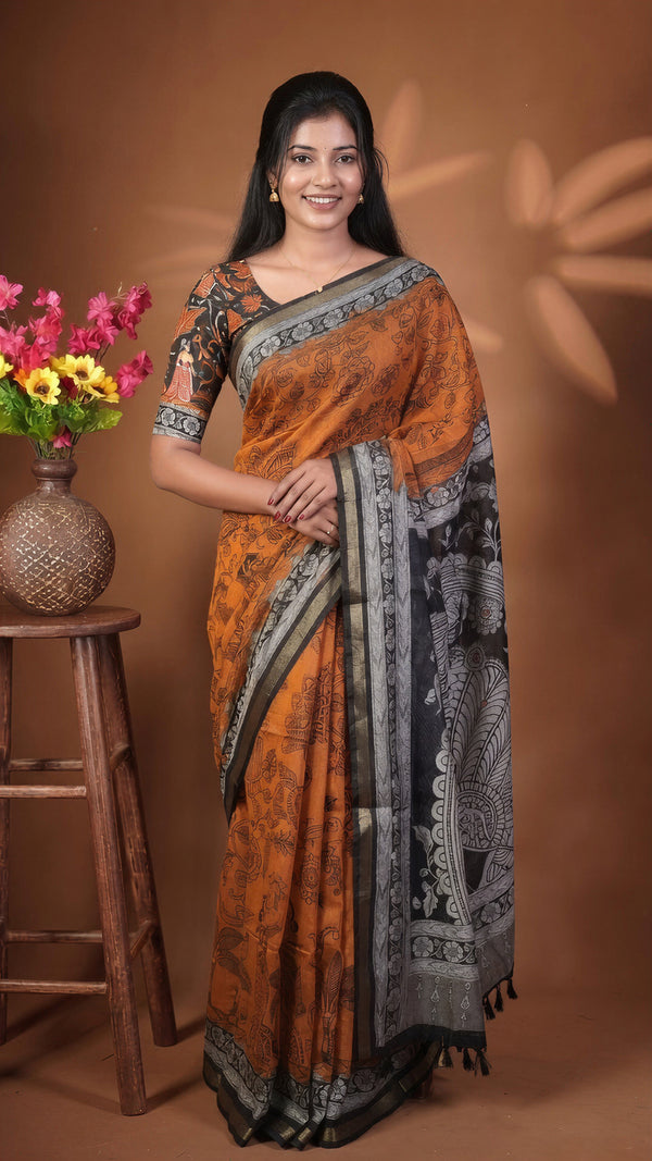 A woman modeling a traditional Indian saree with an orange body, black and gray border patterns, and a matching printed blouse, standing against a warm brown studio backdrop next to a wooden stool with flowers.