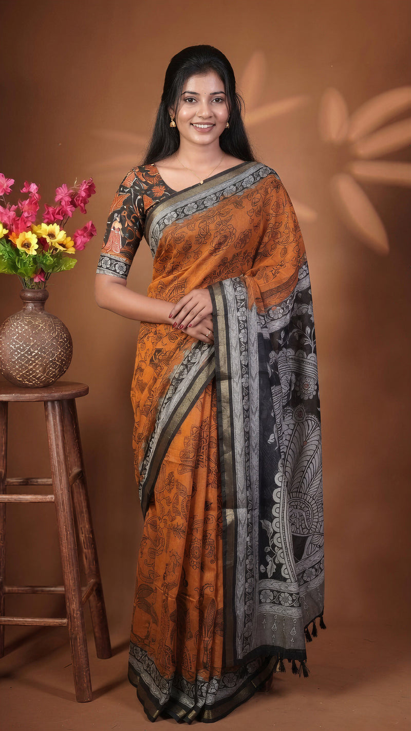 A woman modeling a traditional Indian saree with an orange body, black and gray border patterns, and a matching printed blouse, standing against a warm brown studio backdrop next to a wooden stool with flowers.