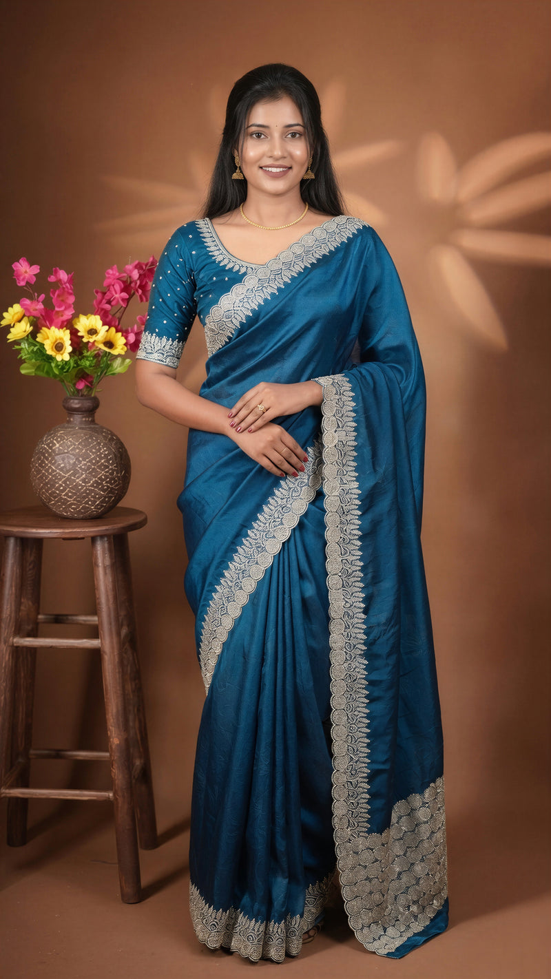 A woman modeling a teal blue saree with intricate silver embroidery on the border and blouse, standing in front of a warm brown backdrop with a wooden stool and flowers on the left.