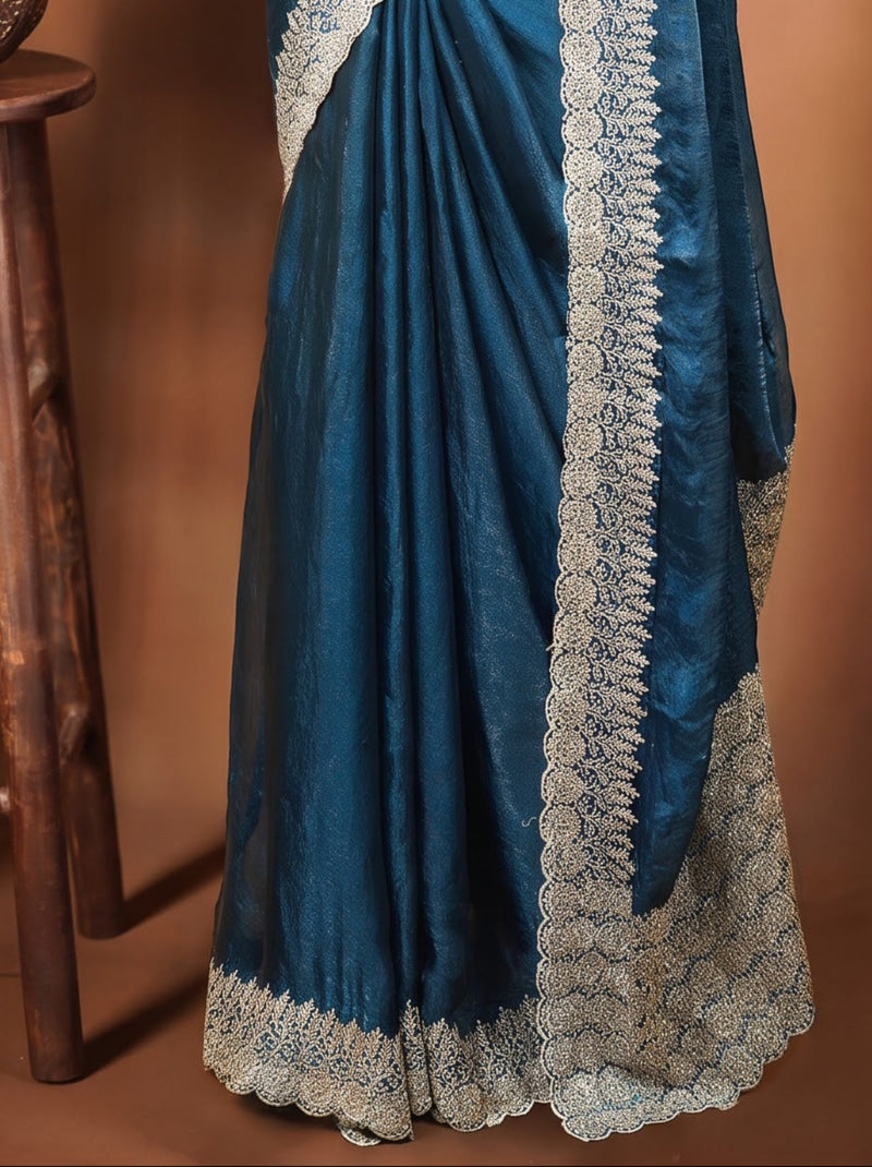 A teal blue silk saree with intricate silver embroidery, displayed on a black mannequin. The mannequin is set against a warm brown backdrop with decorative leaf shadows, and a small wooden stool with a vase of colorful flowers is positioned to the left.