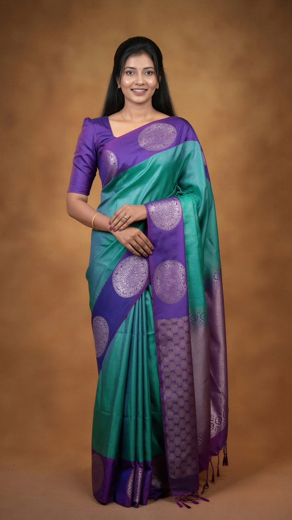 A woman modeling a purple and teal silk saree with metallic circular motifs, set against a solid brown studio backdrop.