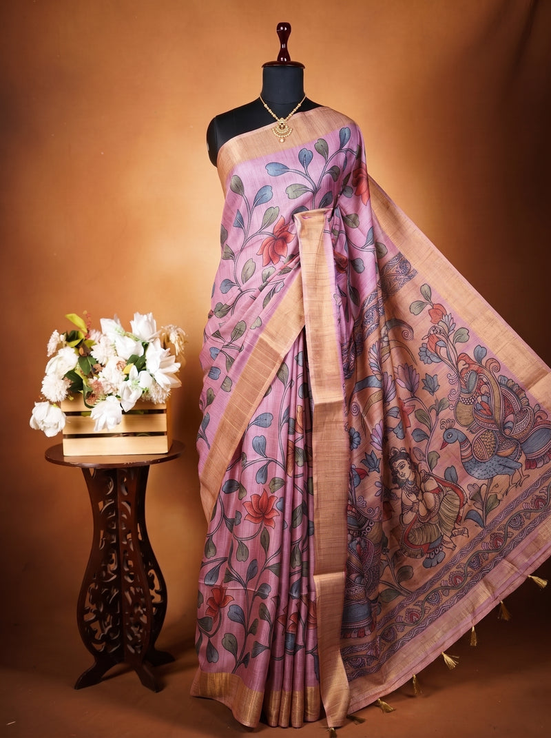A pink and beige printed saree draped on a black mannequin, displayed against a warm brown studio backdrop. A small wooden side table with white flowers is placed to the left.
