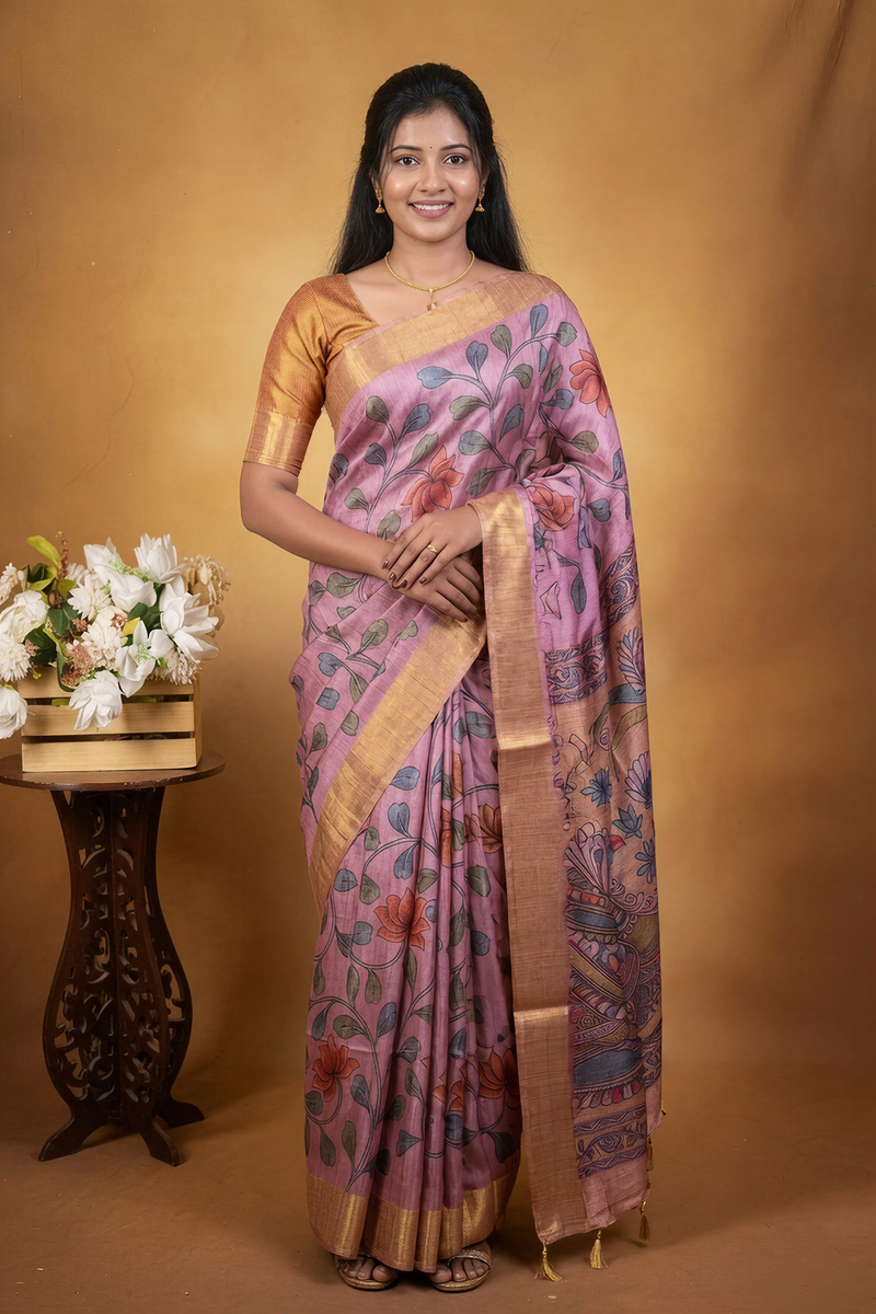 A woman modeling a pink and gold patterned silk saree with floral motifs, standing against a warm brown studio backdrop next to a small table with white flowers.