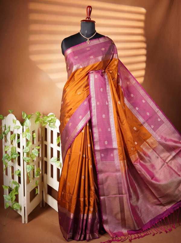 A purple and orange silk saree with a metallic silver border and small white motifs, draped on a black mannequin torso. The mannequin is set against a warm brown background with decorative shadow patterns, a white picket fence, and green vines.