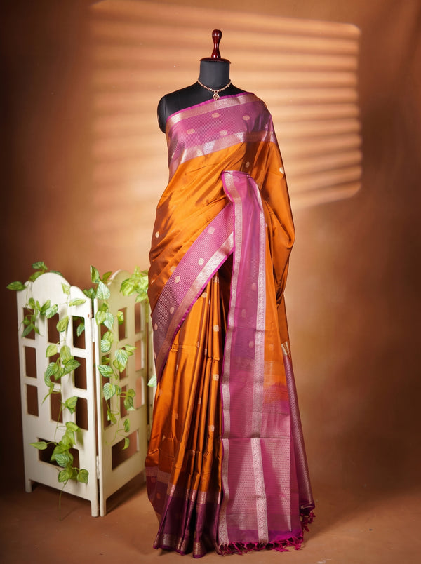 A draped orange and magenta silk saree with white zari patterns, displayed on a black mannequin torso against a warm brown backdrop with decorative leaves on a white stand.