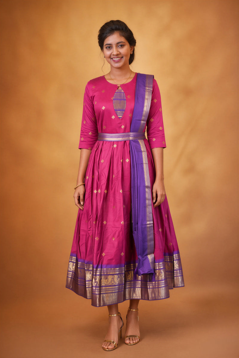 A woman modeling a magenta pink and purple traditional Indian dress with gold zari embroidery, standing against a warm brown studio backdrop.
