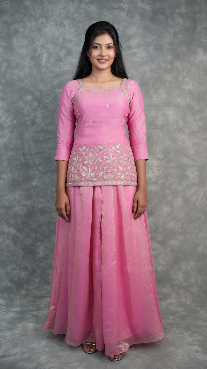 A woman modeling a pink traditional outfit consisting of a three-quarter sleeve kurta with floral embroidery and a matching long skirt, set against a mottled gray studio backdrop.