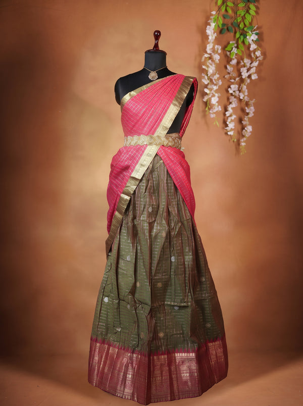 A traditional Indian saree displayed on a black mannequin torso against a warm brown studio backdrop. The saree has a bright pink upper drape (pallu) with a metallic gold border, paired with a dark olive green body featuring gold zari patterns and a matching pink and gold border at the bottom.