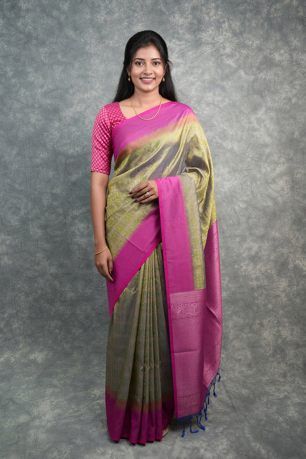 A woman modeling a traditional Indian saree with a vibrant magenta-pink border and blouse, paired with a body fabric that transitions from olive green to gold with subtle zari embroidery, set against a textured gray studio backdrop.