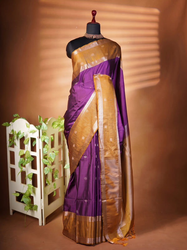 A purple silk saree with a metallic gold border and pallu draped on a black mannequin, set against a warm brown backdrop with a decorative white planter on the left.